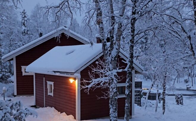 A small red wooden cabin with snow-covered roof and trees stands in a snowy landscape at dusk, with a warm light glowing by the door.