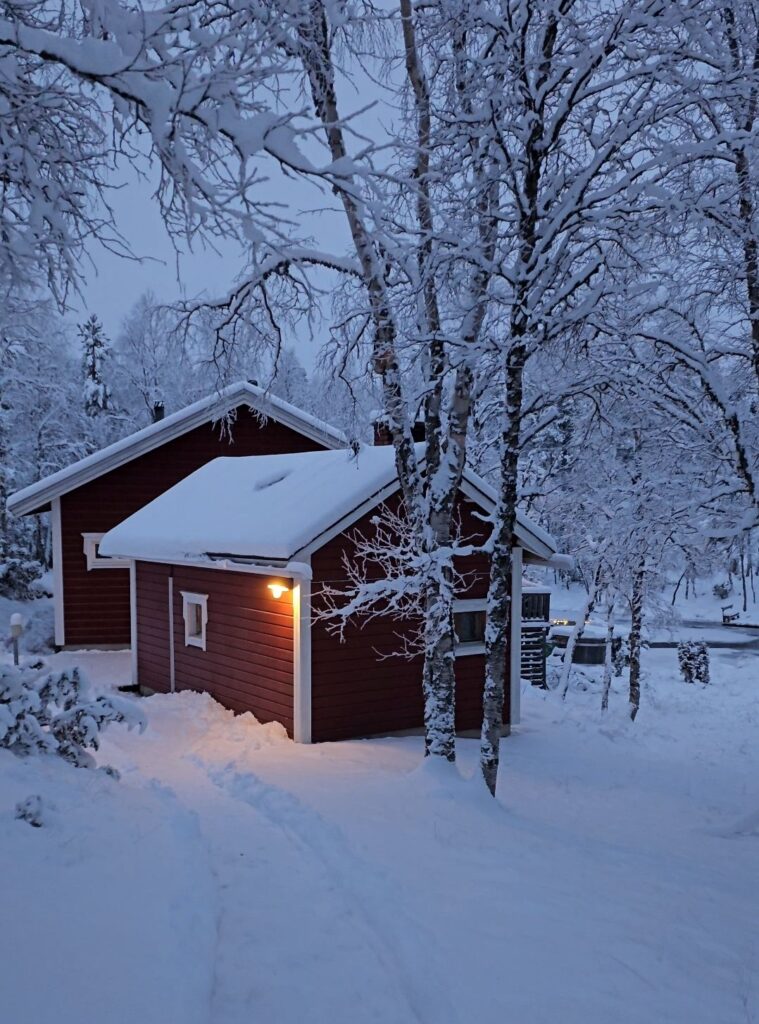 A small red wooden cabin with snow-covered roof and trees stands in a snowy landscape at dusk, with a warm light glowing by the door.