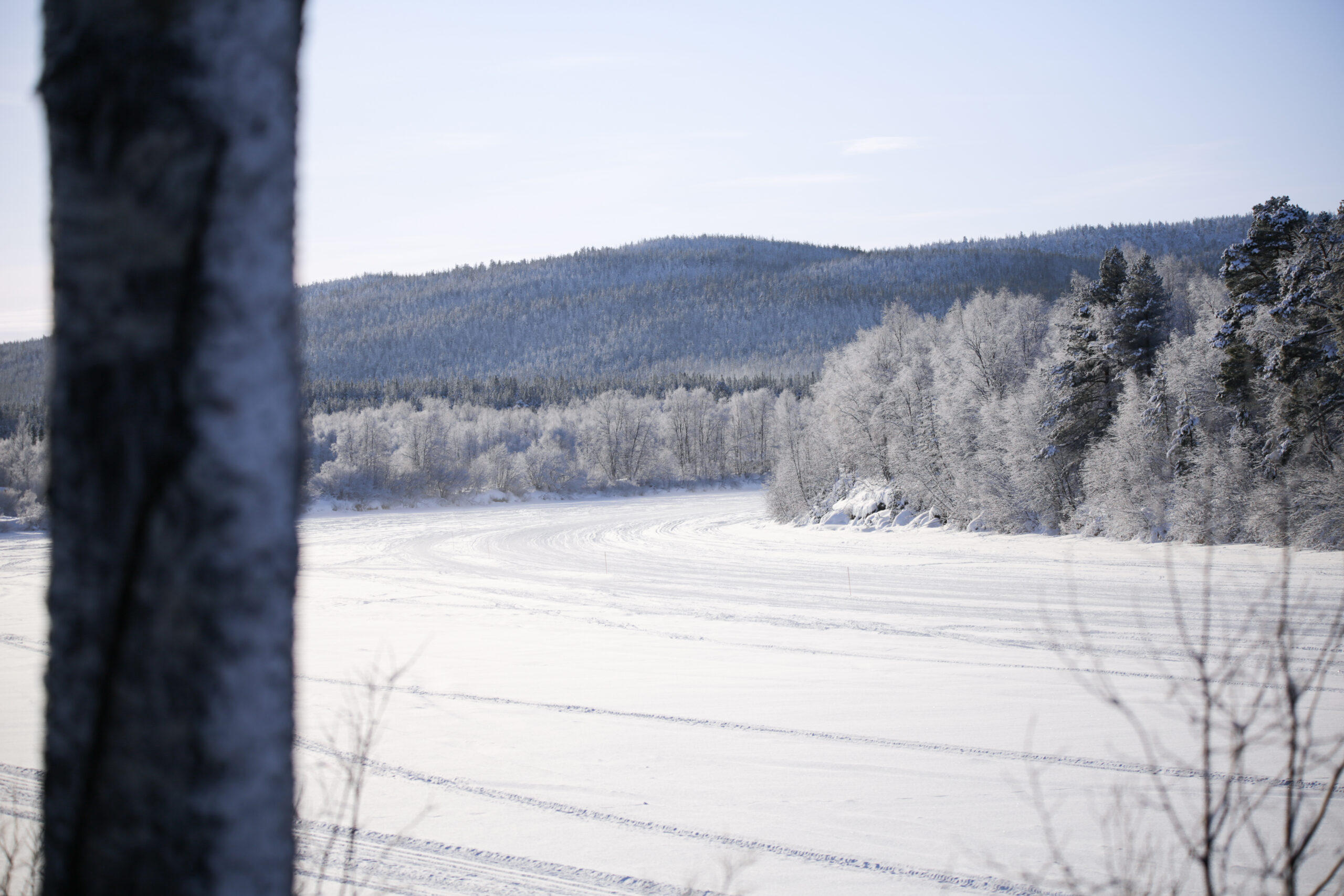 Lumipeitteinen maisema, jossa on huurteiset puut, jäätynyt joki ja kaukana siintävät kukkulat kirkkaan sinisen taivaan alla, tuo mieleen Pohjois-Lapin matkakohteiden rauhallisen kauneuden. Kuvassa näkyy etualalla vasemmalla puolella puunrunko.