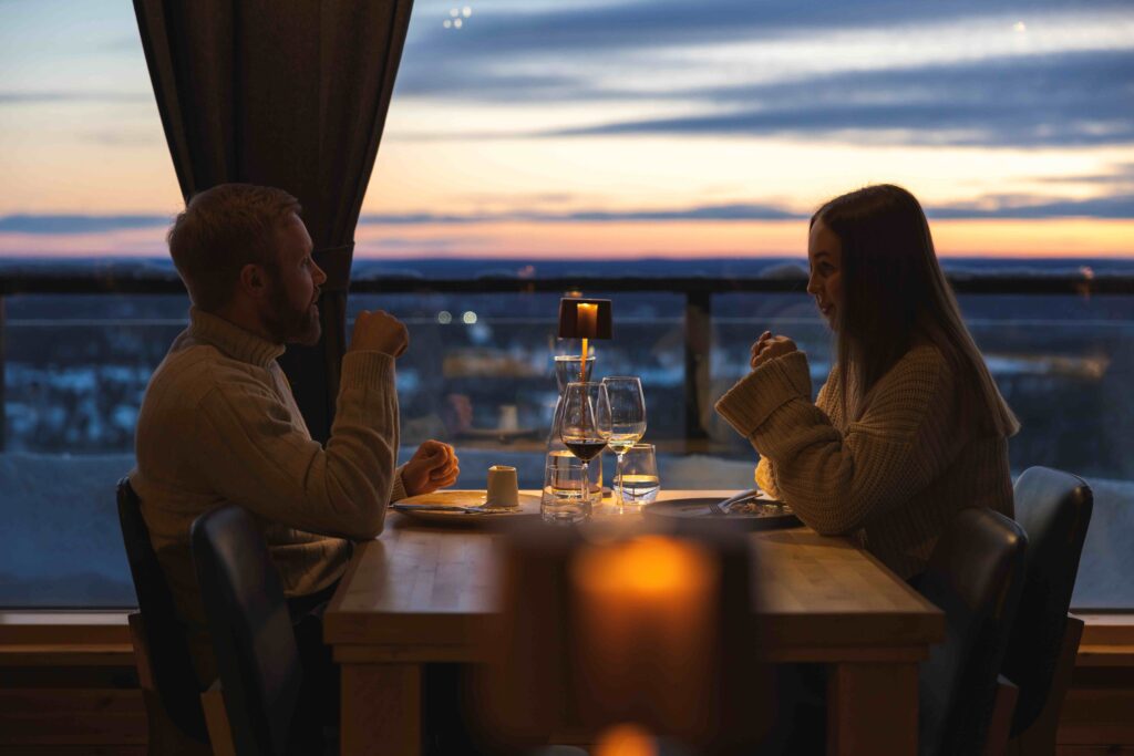 A man and woman sit across from each other at a restaurant table by a window at sunset, engaged in conversation with wine glasses and a candle between them.