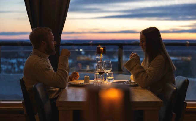 A man and woman sit across from each other at a restaurant table by a window at sunset, engaged in conversation with wine glasses and a candle between them.