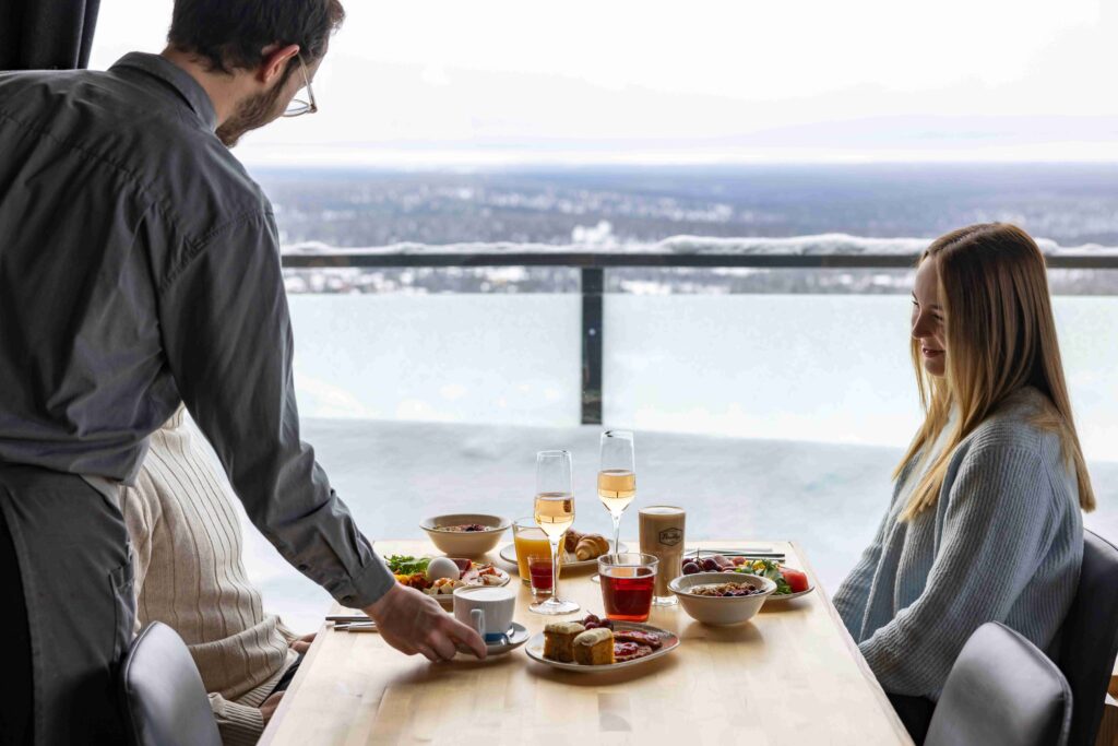 A server places a cup on a table set with food and drinks, as a woman in a sweater smiles. The scene overlooks a snowy landscape through large windows.