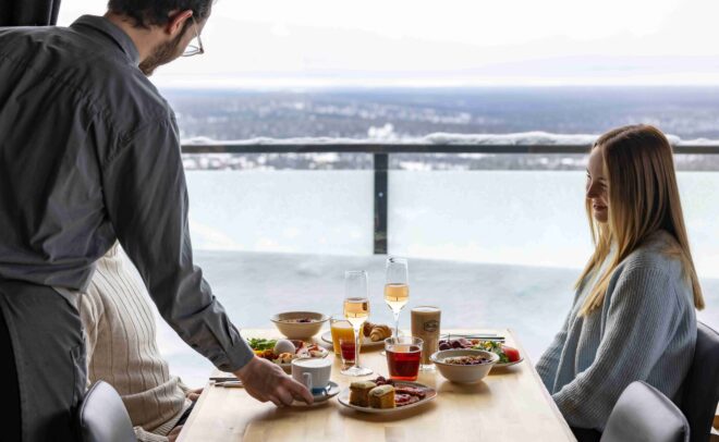 A server places a cup on a table set with food and drinks, as a woman in a sweater smiles. The scene overlooks a snowy landscape through large windows.