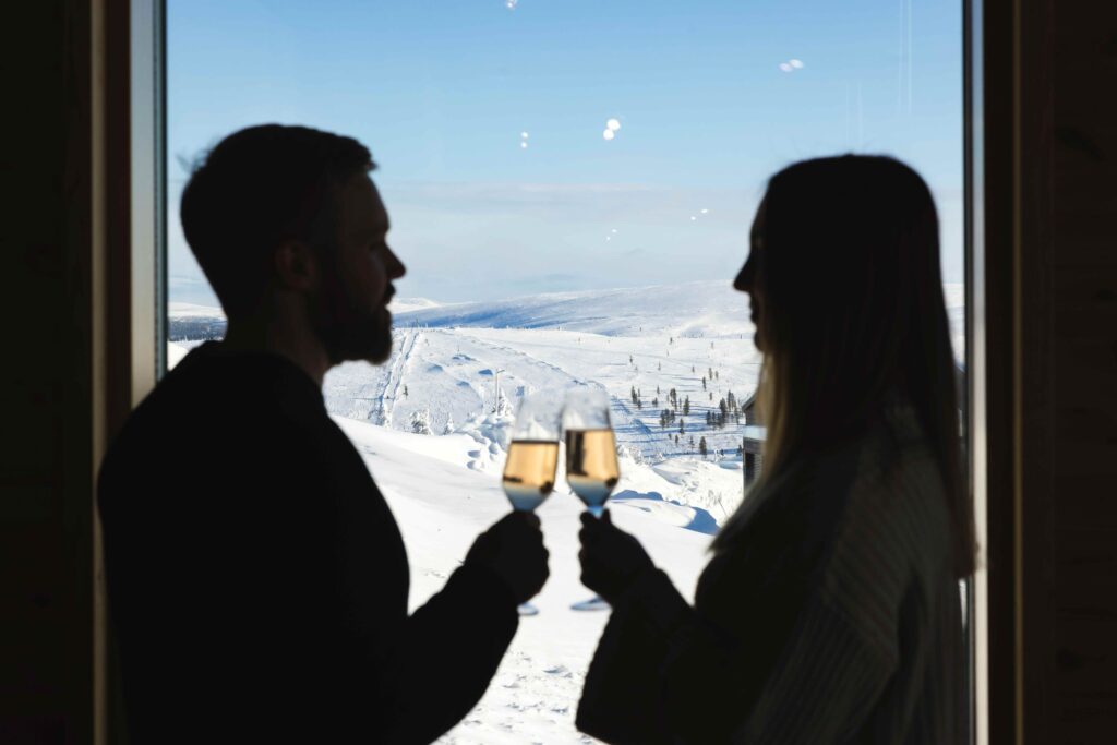 Two people are silhouetted as they clink champagne glasses by a window, with a snowy landscape and clear blue sky visible outside.