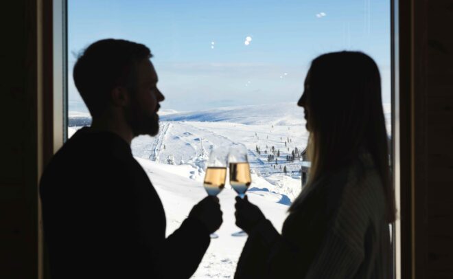 Two people are silhouetted as they clink champagne glasses by a window, with a snowy landscape and clear blue sky visible outside.