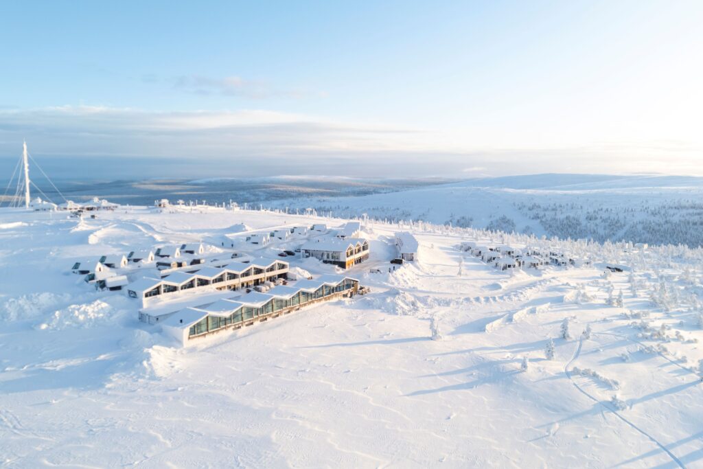 Aerial view of a modern resort complex surrounded by snowy, rolling hills and scattered trees under a clear, bright sky in a winter landscape.