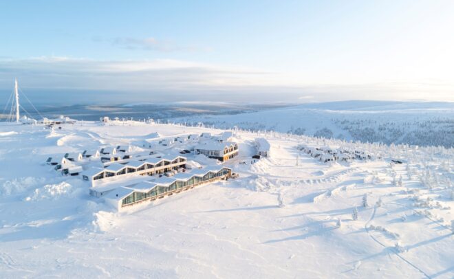 Aerial view of a modern resort complex surrounded by snowy, rolling hills and scattered trees under a clear, bright sky in a winter landscape.