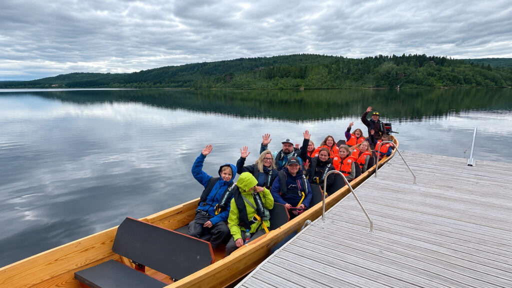 Ryhmä värikkäissä takkeissa pukeutuneita ihmisiä istuu veneessä rauhallisella järvellä, hymyillen ja heiluttaen käsiään, taustalla metsäiset kukkulat ja pilvinen taivas; vene on kiinnitetty puisen laiturin viereen.