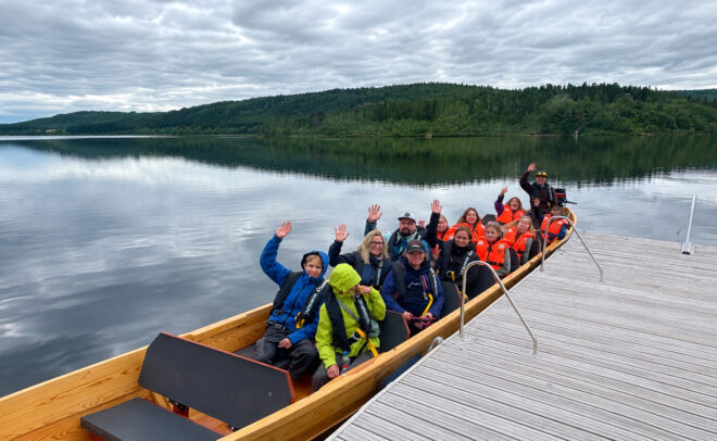 Ryhmä värikkäissä takkeissa pukeutuneita ihmisiä istuu veneessä rauhallisella järvellä, hymyillen ja heiluttaen käsiään, taustalla metsäiset kukkulat ja pilvinen taivas; vene on kiinnitetty puisen laiturin viereen.