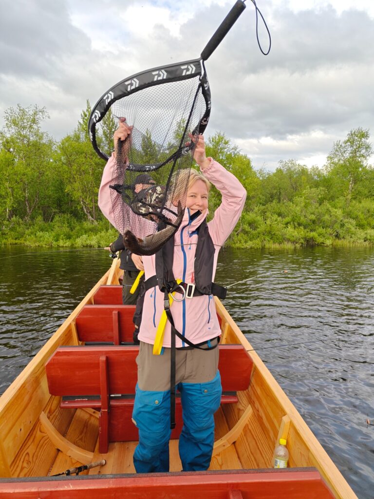 Vaaleanpunaisessa takissa ja sinisissä housuissa oleva henkilö seisoo puuveneessä järvellä, hymyilee ja nostaa ylös kalaverkkoa, jossa on kala. Taustalla näkyy vihreitä puita ja pilvinen taivas.