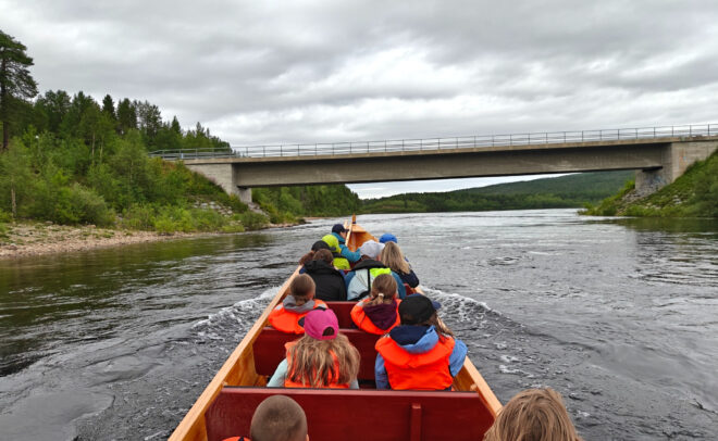 Ryhmä ihmisiä, joilla on yllään oranssit pelastusliivit, matkustaa puuveneellä joella kohti siltaa, ympärillään vihreitä puita ja pilvinen taivas.