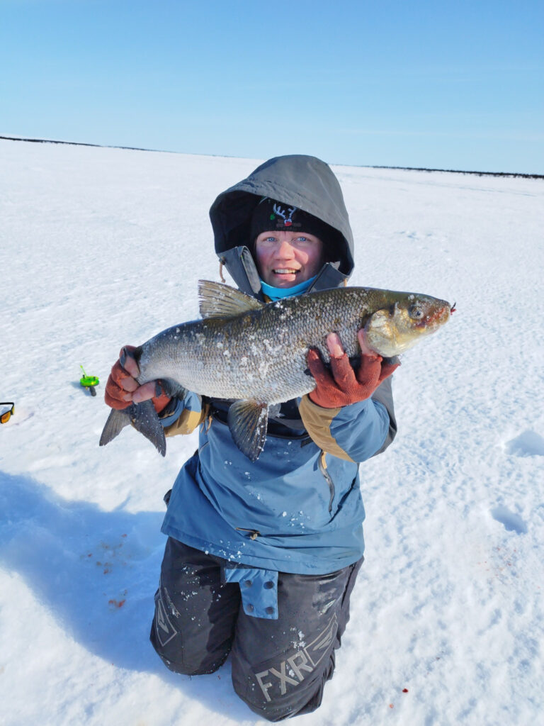 Talvivaatteisiin pukeutunut henkilö seisoo lumella ja jäällä, hymyilee ja pitelee molemmin käsin suurta kalaa. Taivas on kirkas ja sininen, ja maassa lähellä näkyy pieni vihreä kalastuskelan.