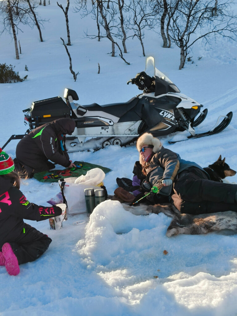 Kolme talvivaatteisiin pukeutunutta henkilöä rentoutuu lumella moottorikelkan vieressä. Yksi heistä makaa koiran kanssa, toinen polvistuu valmistamassa ruokaa tai juomaa, kun taas vaaleanpunaisissa saappaissa oleva lapsi istuu lähellä. Taustalla näkyy lumipeitteisiä puita.