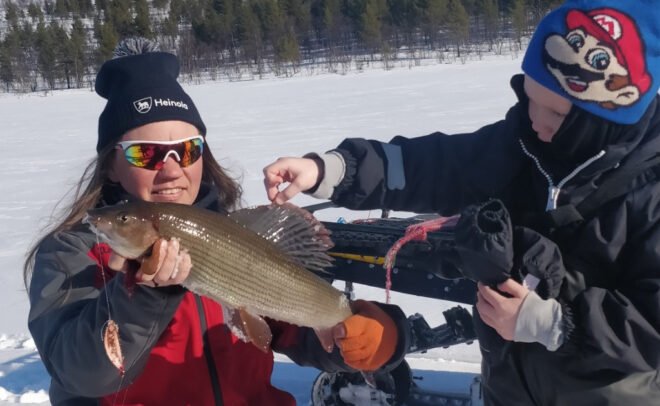 Kaksi talvivaatteisiin pukeutunutta henkilöä poseeraa lumella; toinen pitää kädessään suurta kalaa, kun taas toinen osoittaa sitä sormella. Taustalla näkyy lumisia kukkuloita ja puita kirkkaan sinisen taivaan alla.