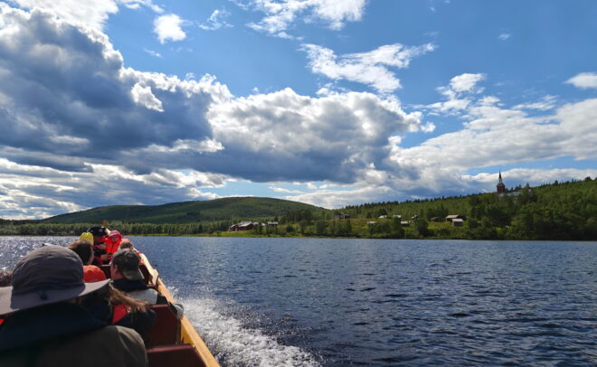 Veneessä olevat ihmiset ajavat järvellä, jonka rannoilla kohoaa metsäisiä kukkuloita ja hajallaan sijaitsevia taloja osittain pilvisen taivaan alla, jättäen jälkeensä aallon.