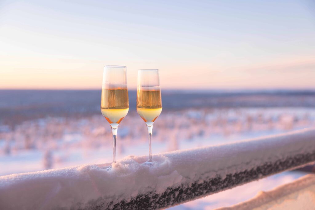 Two champagne glasses filled with sparkling wine rest on a snowy railing, overlooking a serene, snow-covered landscape at sunset.