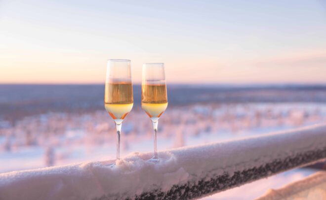 Two champagne glasses filled with sparkling wine rest on a snowy railing, overlooking a serene, snow-covered landscape at sunset.