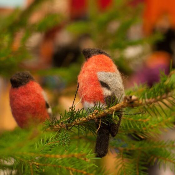 Two felt bird ornaments with red, black, and gray colors perch on green fir tree branches, with a blurred indoor background.