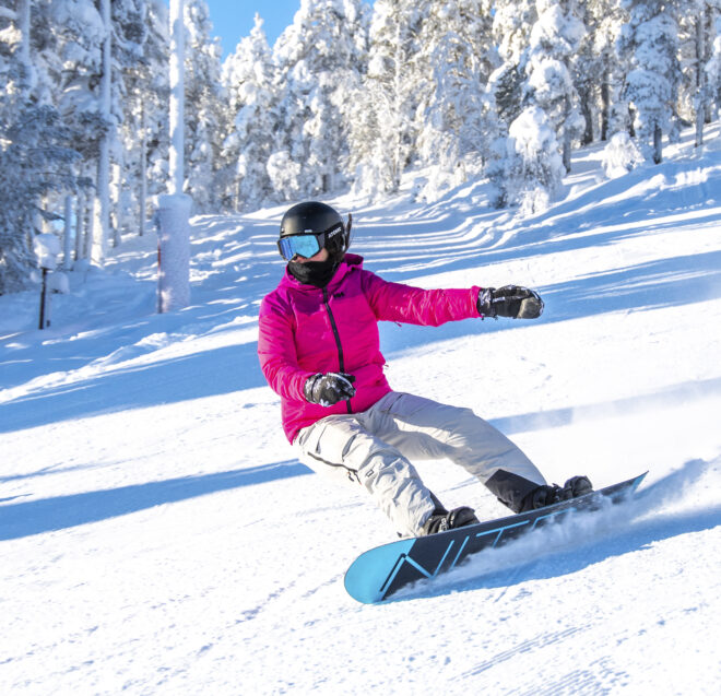 Un snowboardeur, vêtu d'une veste rose vif et d'un casque noir, dévale une pente enneigée bordée de pins chargés de neige.