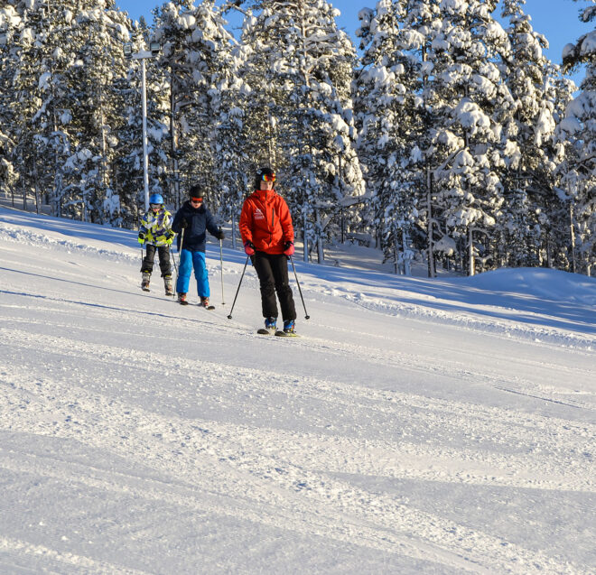 Kolme ihmistä laskee suksilla alas lumipeitteistä rinnettä, jota reunustavat lumiset männyt.