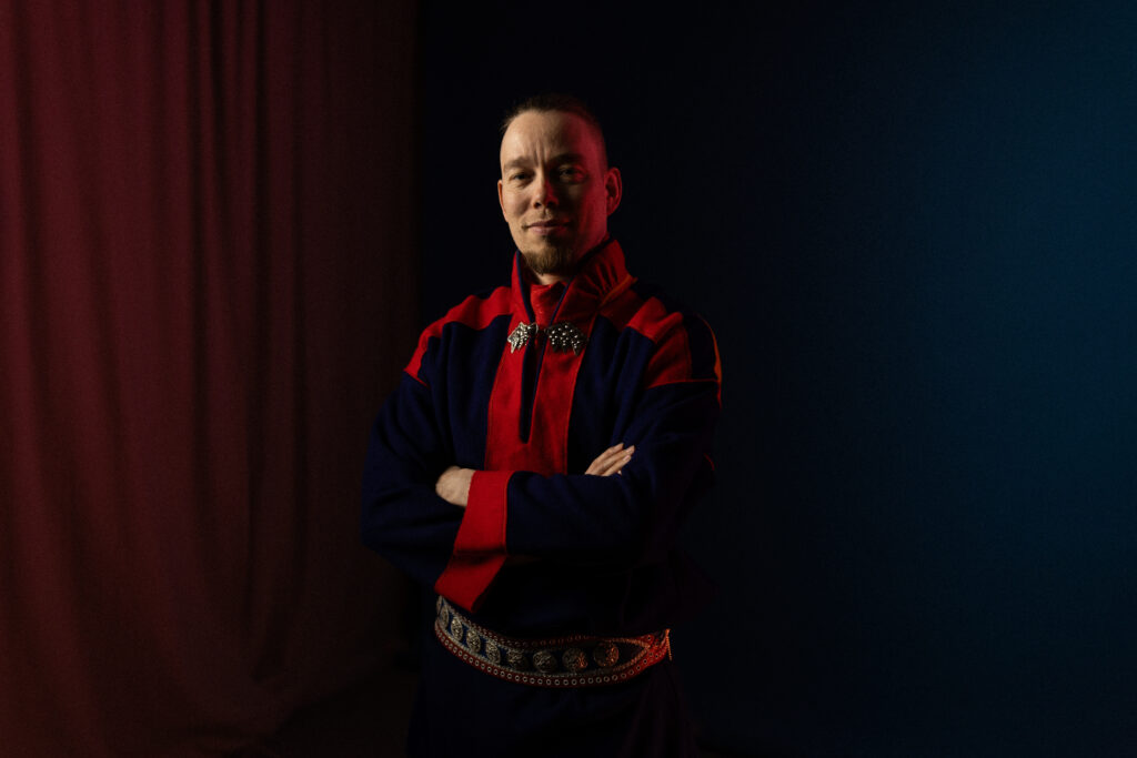Henkilö, jolla on yllään sininen ja punainen perinteinen puku, jossa on koristeelliset soljet, seisoo kädet ristissä ja hymyilee hieman tummansinisen ja viininpunaisen taustan edessä.