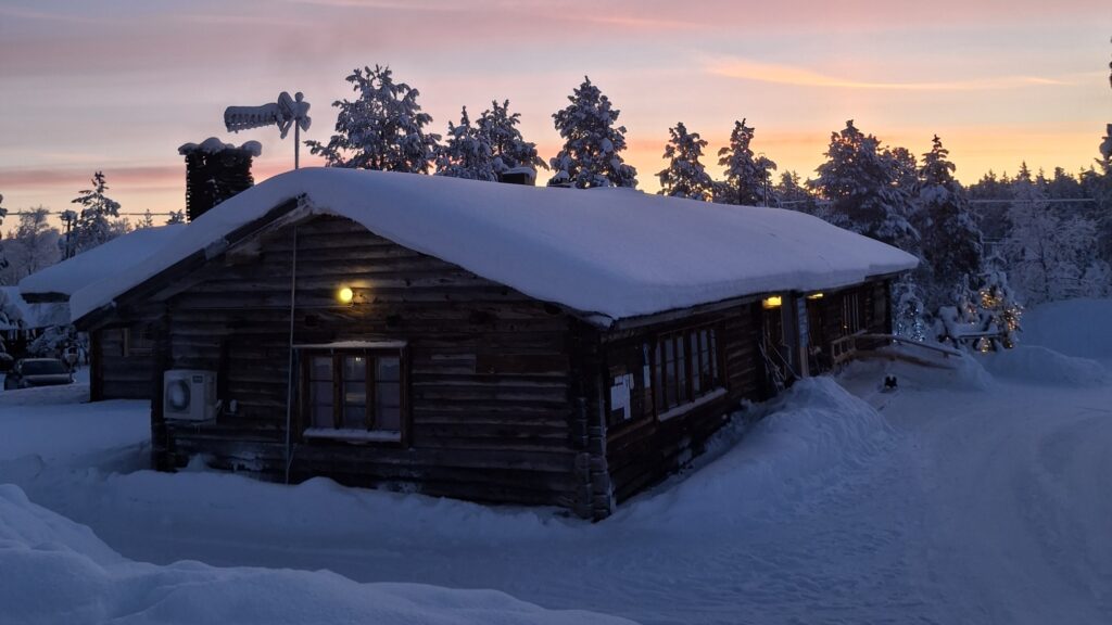 A snow-covered log cabin illuminated by warm exterior lights at twilight, surrounded by snow-laden pine trees.