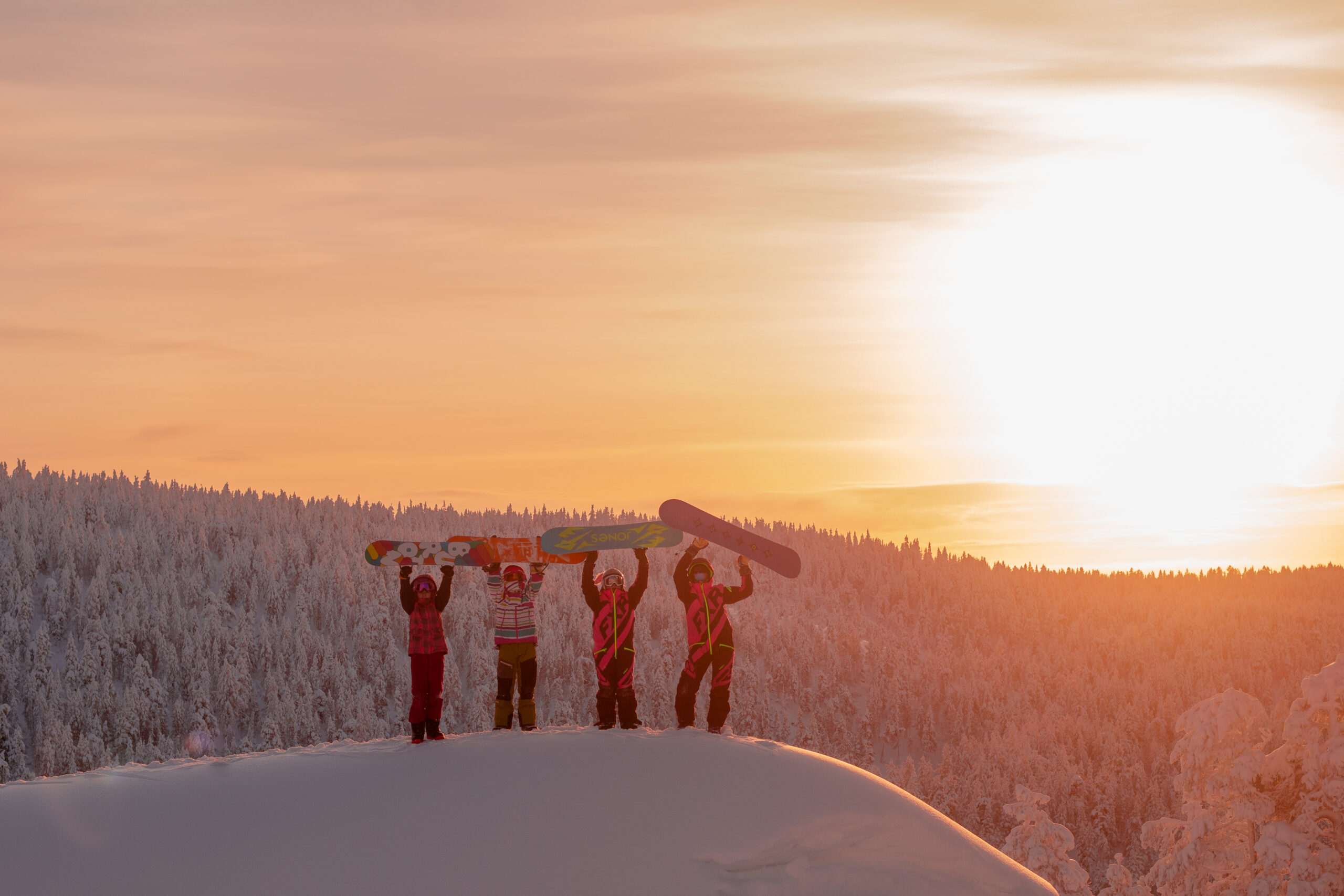 Vier Snowboarder stehen auf einem verschneiten Bergrücken und halten ihre Snowboards vor dem Hintergrund des Sonnenuntergangs über ihre Köpfe.