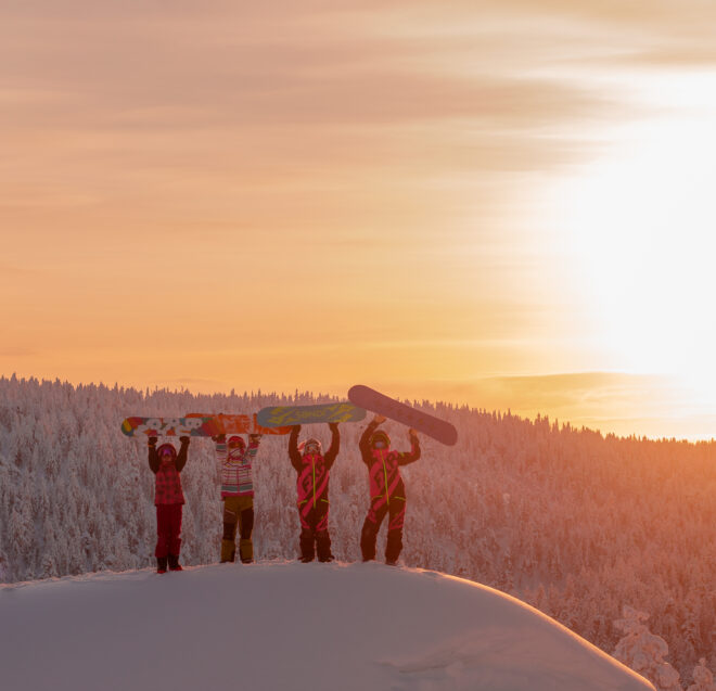 Neljä ihmistä seisoo lumisella harjanteella auringonlaskun aikaan, ja jokainen heistä pitää lumilautaa päänsä yläpuolella.