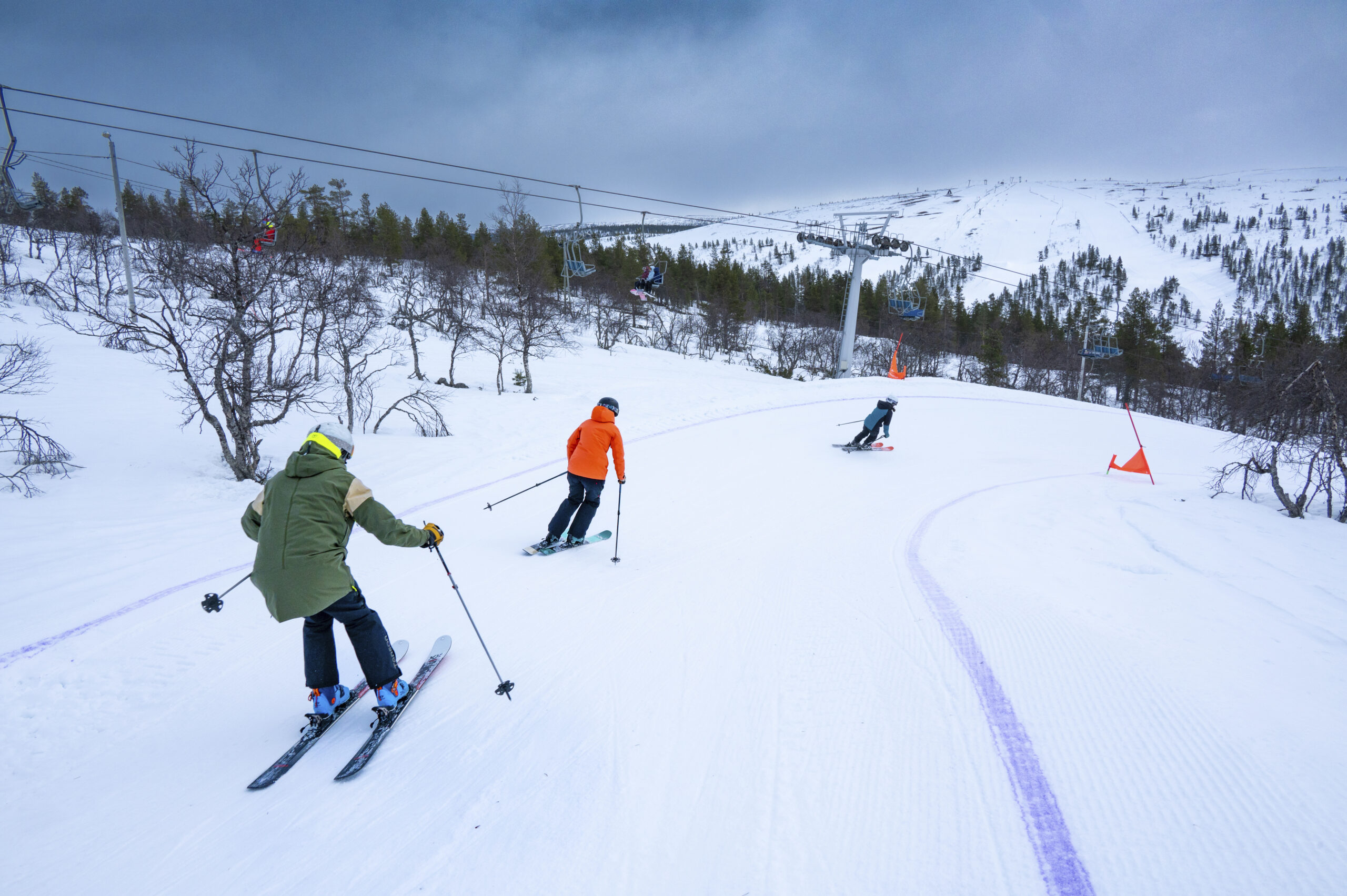 Kolme henkilöä laskee suksilla lumipeitteistä rinnettä alas hiihtohissien ja puiden lähellä pilvisen taivaan alla; rinteen reunalla näkyy oransseja merkkejä.