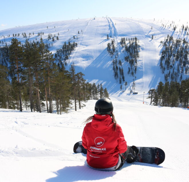 Ein Snowboarder, der eine rote Jacke der Skischule Saariselkä trägt, sitzt auf einem verschneiten Hang und blickt auf eine weit entfernte Skipiste.